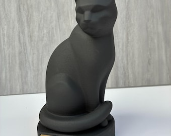 May include: A matte black cat figurine in a seated position, mounted on a black base with a gold-colored rectangular plaque. The plaque features the name Dexter in a script font, followed by the years 2010-2026 in a standard sans-serif font.