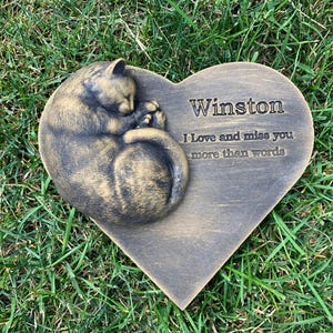 May include: A heart-shaped memorial stone with a sleeping cat sculpture. The stone is engraved with the name "Winston" and the message "I love and miss you more than words".