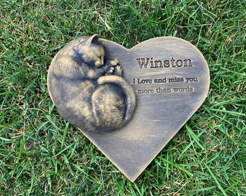May include: A heart-shaped memorial stone with a sleeping cat sculpture. The stone is engraved with the name "Winston" and the message "I love and miss you more than words".