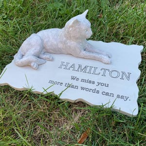 K&ouml;nnte beinhalten: Wei&szlig;er Keramik-Gedenkstein mit einer darauf liegenden Katzenfigur. Der Stein tr&auml;gt die Inschrift "HAMILTON We miss you more than words can say."