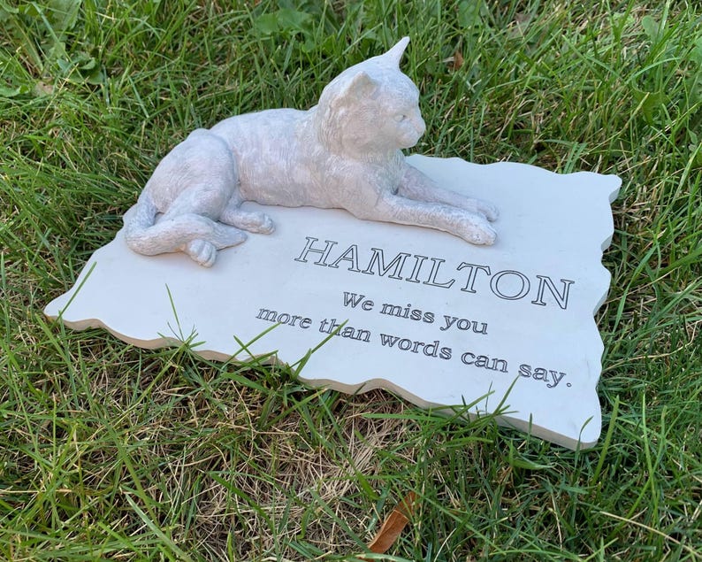 K&ouml;nnte beinhalten: Wei&szlig;er Keramik-Gedenkstein mit einer darauf liegenden Katzenfigur. Der Stein tr&auml;gt die Inschrift "HAMILTON We miss you more than words can say."