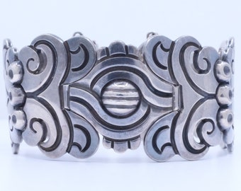 May include: A silver bracelet with a detailed, ornate design featuring swirling patterns and a central circular element. The bracelet is made of polished silver and has a wide, flat band.