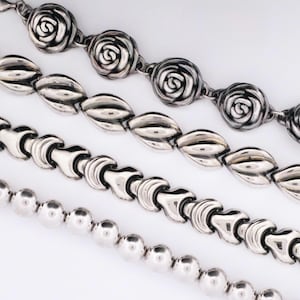 May include: Four silver-toned necklaces with different designs are displayed against a white background. The top necklace features rose-shaped links, followed by an oval-linked chain, a chain with curved links, and a chain of small, round beads.