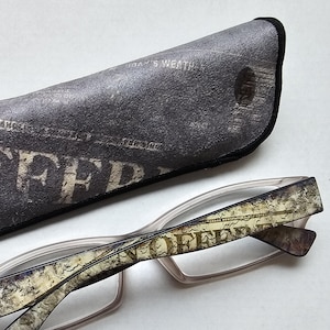 May include: A pair of spectacles with translucent frames and temples featuring a distressed newspaper print pattern that includes the word OFFER. The spectacles are accompanied by a matching triangular case with a dark grey newspaper print.