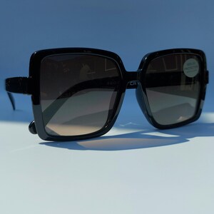 May include: Black square sunglasses with gradient lenses. The sunglasses have a thick frame and a modern design. The lenses transition from a darker shade at the top to a lighter shade at the bottom. The sunglasses are on a light blue surface.