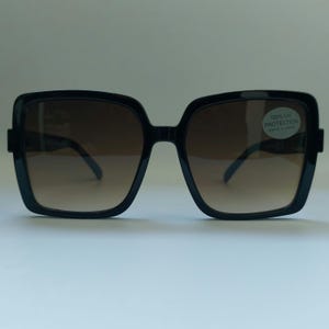 May include: Black square-framed sunglasses with gradient brown lenses. The sunglasses have a thick frame and a small sticker on the right lens that reads "100% UV PROTECTION".