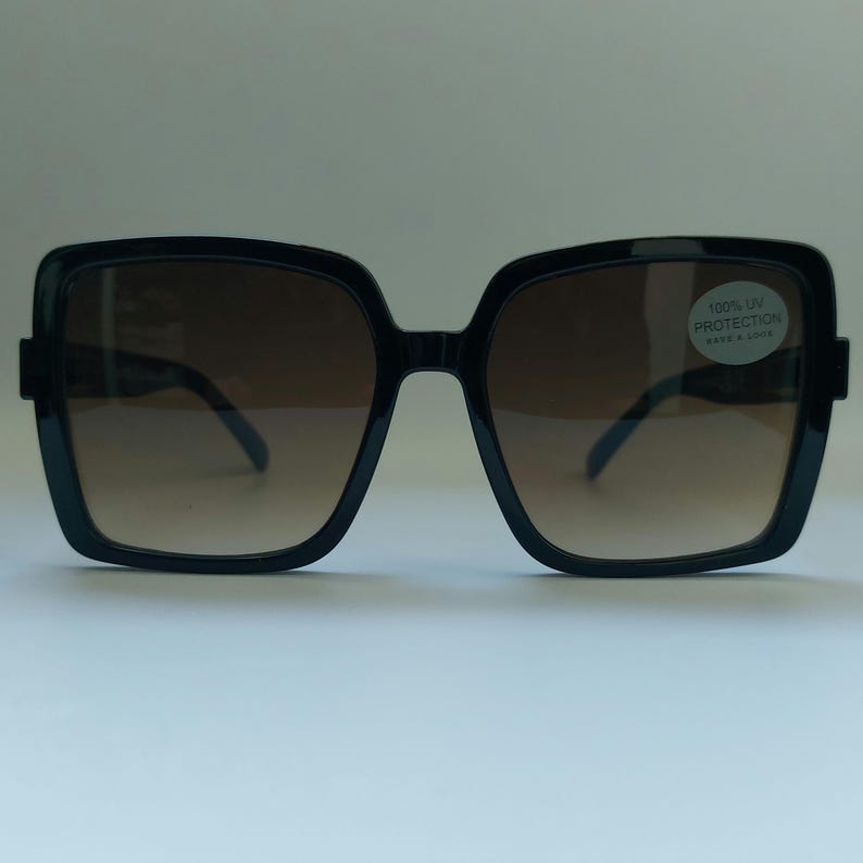May include: Black square sunglasses with brown gradient lenses. The sunglasses have a thick frame and a small sticker on the right lens that reads "100% UV PROTECTION".
