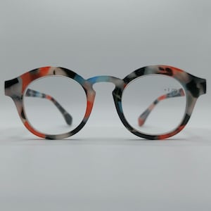 Puede incluir: Gafas de montura redonda con un colorido patrón de rojo, negro, blanco y azul. Las gafas tienen una graduación de +1,00. Las patillas y la montura presentan un diseño moteado. Las lentes son transparentes.