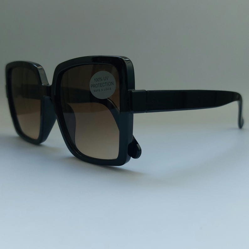 May include: Black square sunglasses with brown gradient lenses. The sunglasses have thick frames and arms. A sticker on the lens reads "100% UV PROTECTION RAYS & LOOK".