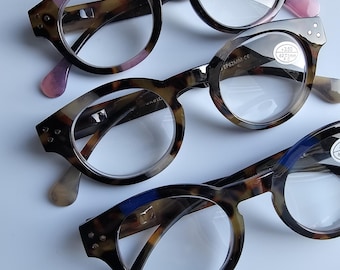 Könnte beinhalten: Drei Brillen mit runden Rahmen im Schildpattmuster. Die Rahmen haben eine dunkelbraune und beige Farbgebung mit rosa und blauen Akzenten an den Bügeln. Jede Brille hat klare Gläser.