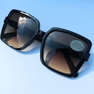 May include: Black square sunglasses with brown gradient lenses. The sunglasses have a sticker that reads "100% UV PROTECTION HAVE A LOOK". The frames are thick and glossy. The sunglasses are set against a light blue background.