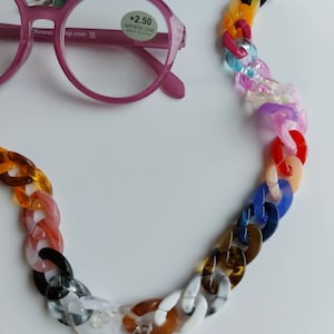 Może przedstawiać: Kolorowy łańcuszek wykonany z plastikowych ogniw w różnych kolorach, w tym czerwonym, pomarańczowym, żółtym, zielonym, niebieskim i fioletowym. Łańcuszek jest przeznaczony do trzymania okularów. Łańcuszek leży na białym tle.