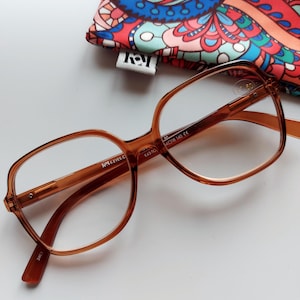 May include: Brown plastic framed glasses with clear lenses. The glasses have the text "Ray-Ban" on the side of the frame and "52 143T C2 54D18 140 CE" on the right temple.