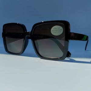 May include: Black square sunglasses with dark gradient lenses. The sunglasses have thick, glossy black frames and arms. A sticker on the lens reads "100% UV PROTECTION".