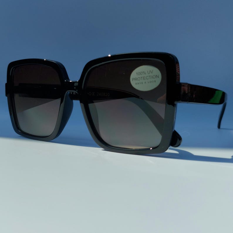 May include: Black square sunglasses with dark gradient lenses. The sunglasses have thick, glossy black frames and arms. A sticker on the lens reads "100% UV PROTECTION".