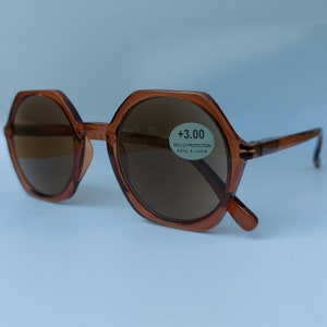 Könnte beinhalten: Braune, achteckige Sonnenbrille mit braunen Gläsern. Die Sonnenbrille hat einen Aufkleber auf dem rechten Glas, auf dem " +3.00 100% UV Protection Have a Look" steht.