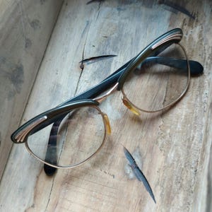 May include: Vintage eyeglasses with a dark green and gold frame. The glasses have a classic design with a thin gold rim around the lenses and a decorative detail on the temples. The frame rests on a wooden surface.