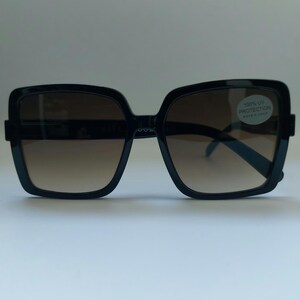 May include: Black square sunglasses with brown gradient lenses. The sunglasses have a thick frame and a sticker that reads "100% UV Protection". The lenses transition from a darker shade at the top to a lighter shade at the bottom.