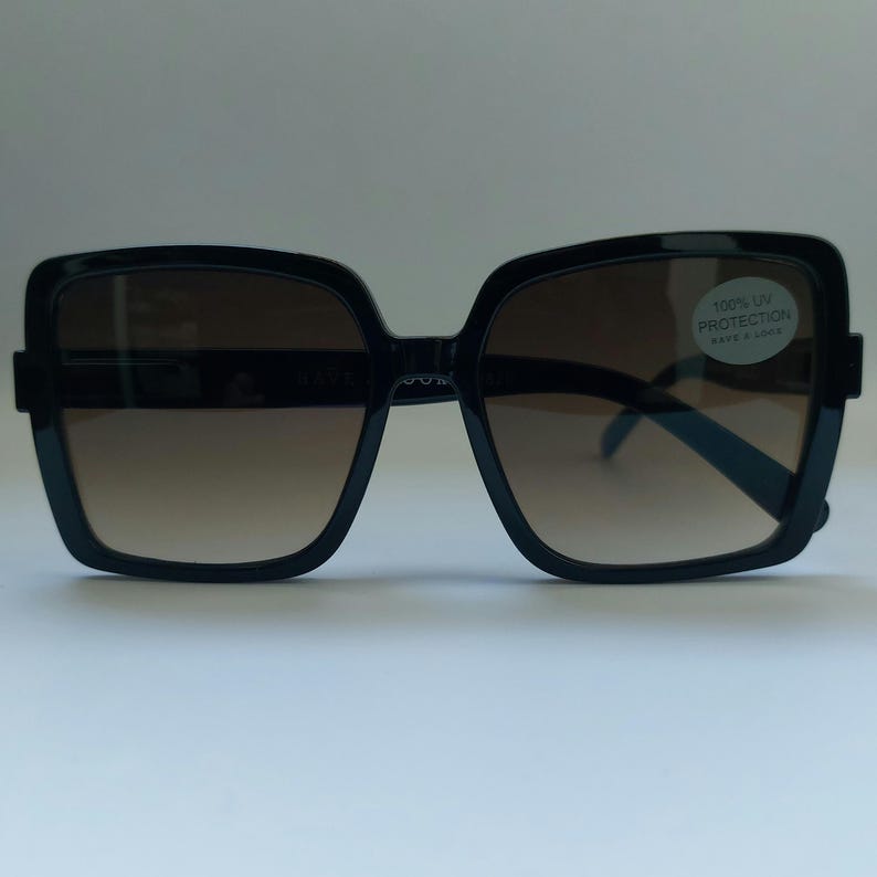May include: Black square sunglasses with brown gradient lenses. The sunglasses have a thick frame and a sticker that reads "100% UV Protection". The lenses transition from a darker shade at the top to a lighter shade at the bottom.