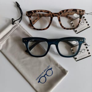 May include: Two pairs of reading glasses with a brown and blue frame. The glasses are on a white surface with a white pouch. The pouch has the text "Hoeksche Brillen" and a pair of glasses logo.