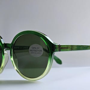 Op de afbeelding: Ronde zonnebril met een doorschijnend groen montuur dat naar onderen toe vervaagt in een lichtere limoentint. De donkere glazen zijn voorzien van een ronde sticker met de zwarte tekst 100% UV PROTECTION HAVE A LOOK op een grijze achtergrond.