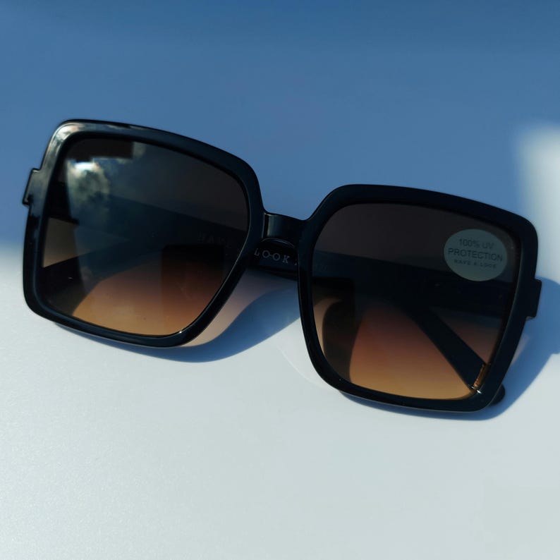 May include: Black square-framed sunglasses with gradient brown lenses. The sunglasses have a thick frame and a small sticker that reads "100% UV PROTECTION". The sunglasses are on a white surface.