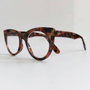 May include: Cat-eye style eyeglasses with a tortoiseshell pattern. The frames are a mix of brown and tan with a shiny surface. The lenses are clear, and the right lens has the text "+1.00". The brand name "Key" is visible on the right temple.