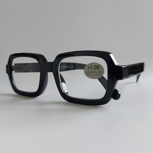 Puede incluir: Gafas de lectura rectangulares negras con una etiqueta de lente asférica +1.00. Las gafas tienen una montura negra y lentes transparentes.