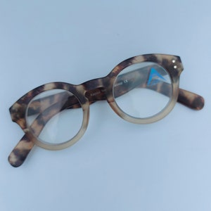 May include: Tortoiseshell reading glasses with a brown and black pattern. The glasses have a round frame and clear lenses. The text "Kapsul for aloop CE" is printed on the frame.