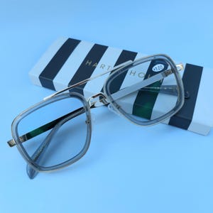 Könnte beinhalten: Silberne Brille mit blauen getönten Gläsern. Die Brille liegt auf einem schwarz-weiß gestreiften Karton mit dem Aufdruck "HART & HOU". Der Karton hat auch einen Aufkleber mit dem Text " +1.00" darauf.
