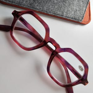 Può includere: Un paio di occhiali da lettura bordeaux e rosa con gradazione +2,50, caratterizzati da una montatura geometrica unica. Gli occhiali sono abbinati a un astuccio in feltro grigio con un logo.