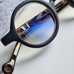 Pode incluir: Close-up de óculos pretos com armação redonda e lentes transparentes. As hastes têm um padrão tartaruga e o texto "in France +1.50". A marca "K-EYES.CO" é visível na armação. Os óculos estão sobre uma superfície branca.