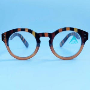 May include: Round brown and black patterned eyeglasses with clear lenses. The right lens has a small green sticker with the text " +1.50" on it.