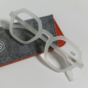 May include: Light grey eyeglasses with a distinctive geometric frame. The glasses are placed on a grey felt case with an orange interior. The lens strength is +2.00.
