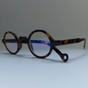 Puede incluir: Gafas redondas con un patrón de carey. Las monturas son marrones y negras con lentes transparentes. Las lentes tienen un tinte azul y la lente derecha tiene la marca "+1.50".