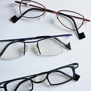 May include: Three pairs of spectacles with distinct frames. The top pair has thin red metal rims. The middle pair features a blue semi-rimless design. The bottom pair has solid black rectangular frames. Each pair includes black temple tips.