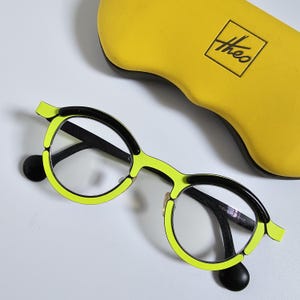 May include: A pair of eyeglasses with a neon yellow frame and black accents. The round lenses are set in a unique design, with a matching yellow case featuring the brand name "theo". The glasses have a modern, stylish look.