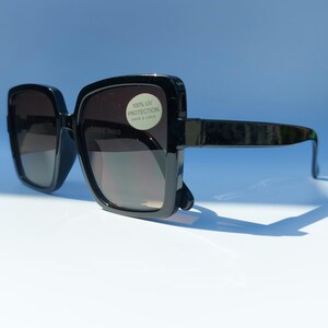 May include: Black square sunglasses with dark lenses. The sunglasses have thick, glossy black frames and arms. A white sticker on the lens reads "100% UV PROTECTION". The sunglasses are set against a light blue background.