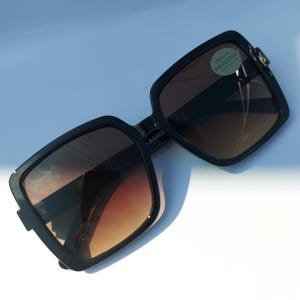 May include: Black square-framed sunglasses with gradient brown lenses. The sunglasses have a modern design with a subtle, angular shape. A small sticker on the lens reads "100% UV PROTECTION".