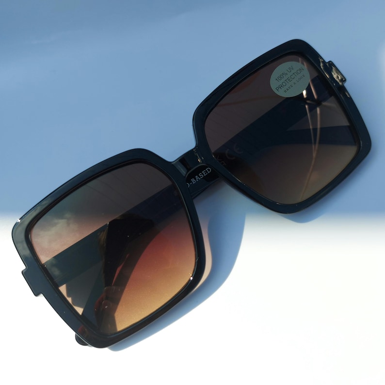 May include: Black square-framed sunglasses with gradient brown lenses. The sunglasses have a modern design with a subtle, angular shape. A small sticker on the lens reads "100% UV PROTECTION".