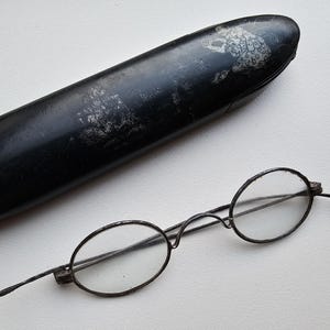 Puede incluir: Gafas antiguas de forma ovalada con una montura fina de metal oscuro y lentes transparentes. Junto a ellas, un estuche largo, negro y ovalado con un diseño descolorido. Las gafas y el estuche están sobre una superficie blanca.