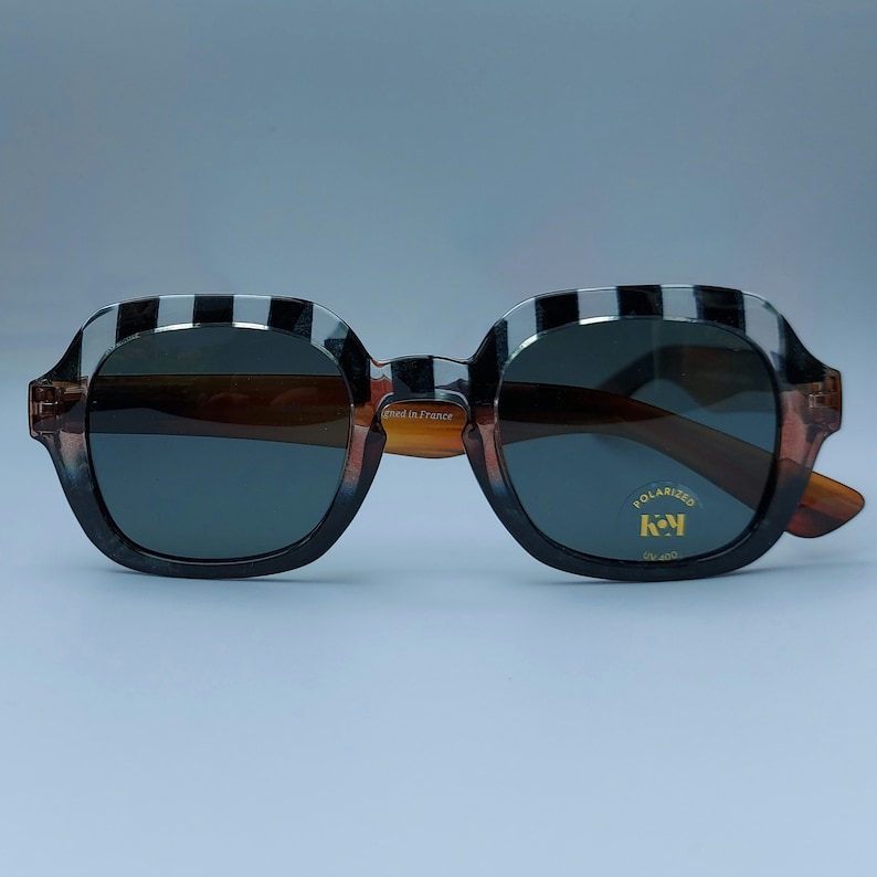 Może przedstawiać: Czarno-białe okulary przeciwsłoneczne w paski z brązowymi szkłami. Okulary mają złoty napis "Made in France" i złotą etykietę z napisem "Polarized K&Y UV 400".