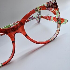 May include: Cat-eye glasses featuring a translucent red frame with a vibrant floral pattern on the inner rim. The lenses are clear and include the printed text 1.50. The design showcases a sharp, pointed upper corner and rounded lower edges.