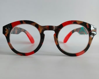 Könnte beinhalten: Runde Lesebrille mit einem schwarzen und roten Schildpattgestell. Die Brille hat klare Gläser und ein kleines blaues Dreieckslogo auf der rechten Linse.