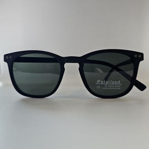 Op de afbeelding: Zwarte zonnebril met een rond montuur en donkere glazen. De zonnebril heeft een matte afwerking en een kleine sticker met de tekst "Polarized" en "UV400 Protection".
