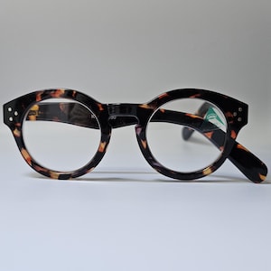 May include: Round-framed spectacles with a tortoiseshell pattern. The frames are dark brown with amber and black accents. The lenses are clear, and the spectacles have a small tag on the right lens. The arms are black.