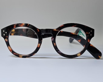Könnte beinhalten: Rundrandbrille mit Schildpattmuster. Die Fassungen sind dunkelbraun mit bernsteinfarbenen und schwarzen Akzenten. Die Gläser sind klar, und die Brille hat ein kleines Etikett auf dem rechten Glas. Die Bügel sind schwarz.