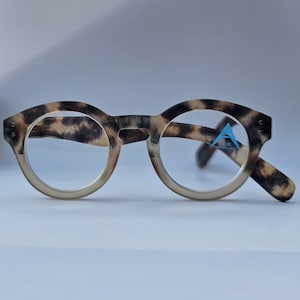 May include: Round reading glasses featuring a brown and tan tortoiseshell pattern on the frames. The right lens displays a small blue triangular sticker with the text +3.00 ASYMMETRIC LENS. The glasses have a thick, sturdy frame design.