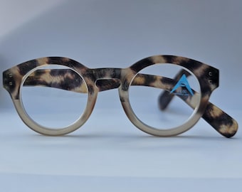 Könnte beinhalten: Runde Lesebrille mit braun-beigem Schildpattmuster auf dem Rahmen. Das rechte Glas zeigt einen kleinen blauen dreieckigen Aufkleber mit dem Text +3.00 ASYMMETRIC LENS. Die Brille hat ein dickes und robustes Rahmendesign.