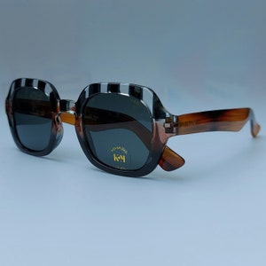 Może przedstawiać: Okulary przeciwsłoneczne w czarno-białe paski z brązowymi oprawkami typu "ż&oacute;łwiowy". Soczewki są ciemne i spolaryzowane. Okulary przeciwsłoneczne mają logo "Koy" na prawej soczewce.